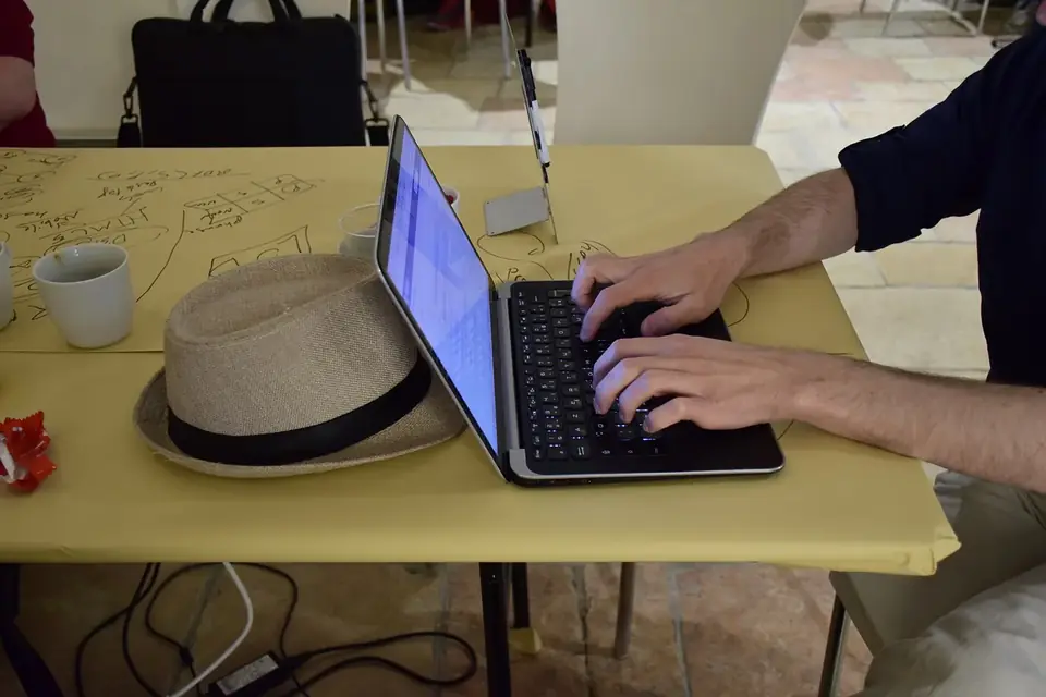 Des mains tapent sur le clavier d'un ordinateur portable posé sur une table jaune, à côté d'un chapeau de paille et de feuilles griffonnées